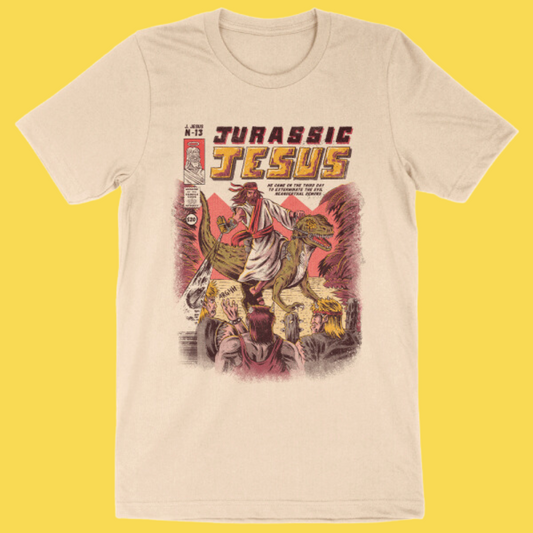 Cream graphic tee featuring 'Jurassic Jesus' vintage comic book style design with dinosaurs and religious imagery on yellow