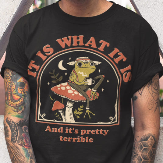 Black graphic tee featuring a whimsical frog on a mushroom holding a coffee cup with text 'It Is What It Is' and 'And it's