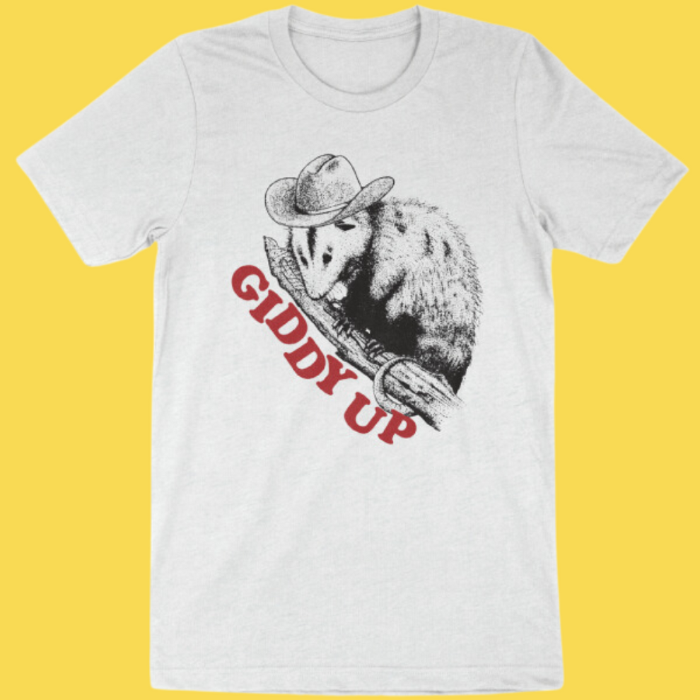 White graphic tee featuring a cowboy opossum holding a rifle with 'Giddy Up' text in red against a yellow background.
