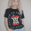 A woman wearing a black t-shirt of of an apple core with seeds that look like a skull and text that reads rotten to the core and bad apple