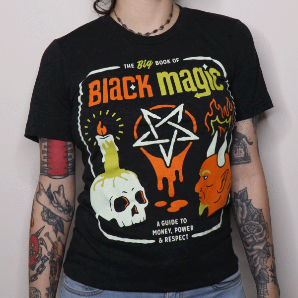 A woman wearing a black t-shirt that reads the big book of black magic: a guide to money, power, and respect with icons of a pentagram, devil head, and skull with a candle burning on top of it