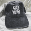 A charcoal grey, unstructured baseball cap with white embroidered text that reads stay weird