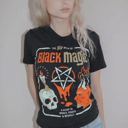A woman wearing a black t-shirt that reads the big book of black magic: a guide to money, power, and respect with icons of a pentagram, devil head, and skull with a candle burning on top of it