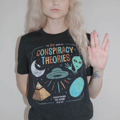 A woman wearing a black graphic tee that reads the big book of conspiracy theories, everything you know is a lie with icons of a UFO space ship, alien, flat earth, and pyramid with an eye