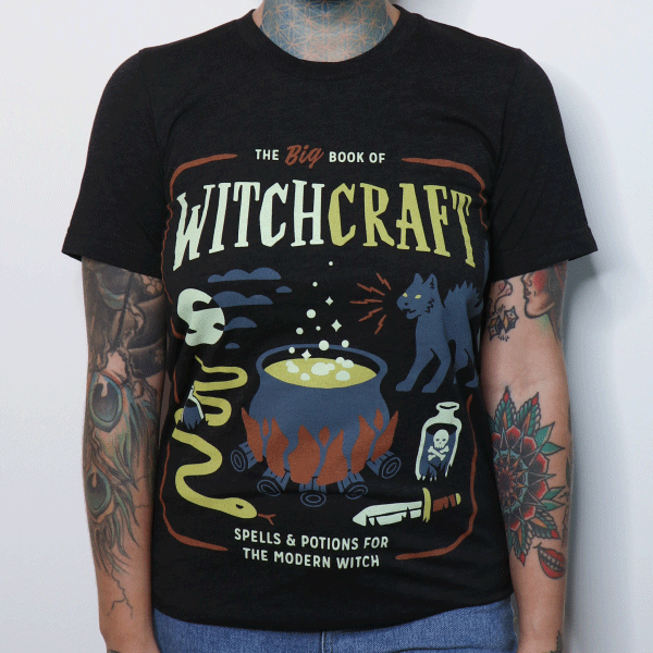 A woman wearing a black graphic tee that reads the big book of witchcraft, spells and potions for the modern witch with icons of a cauldron, black cat, poison bottle, and dagger