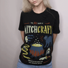 A woman wearing a black graphic tee that reads the big book of witchcraft, spells and potions for the modern witch with icons of a cauldron, black cat, poison bottle, and dagger
